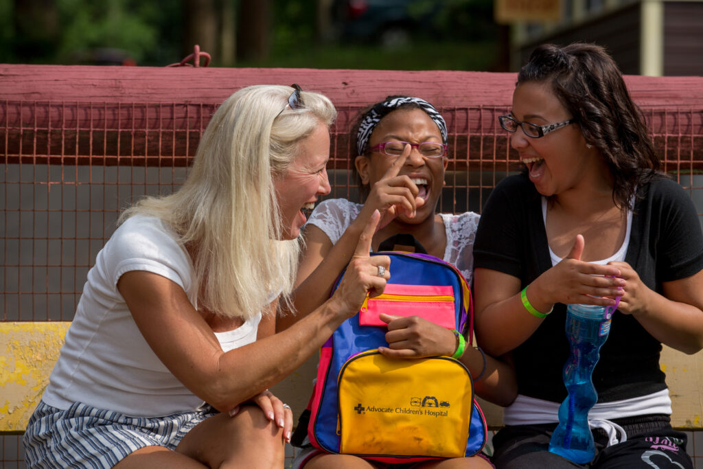 SeriousFun Ambassador Clea Newman laughs with two campers while sitting on a bench at camp.  